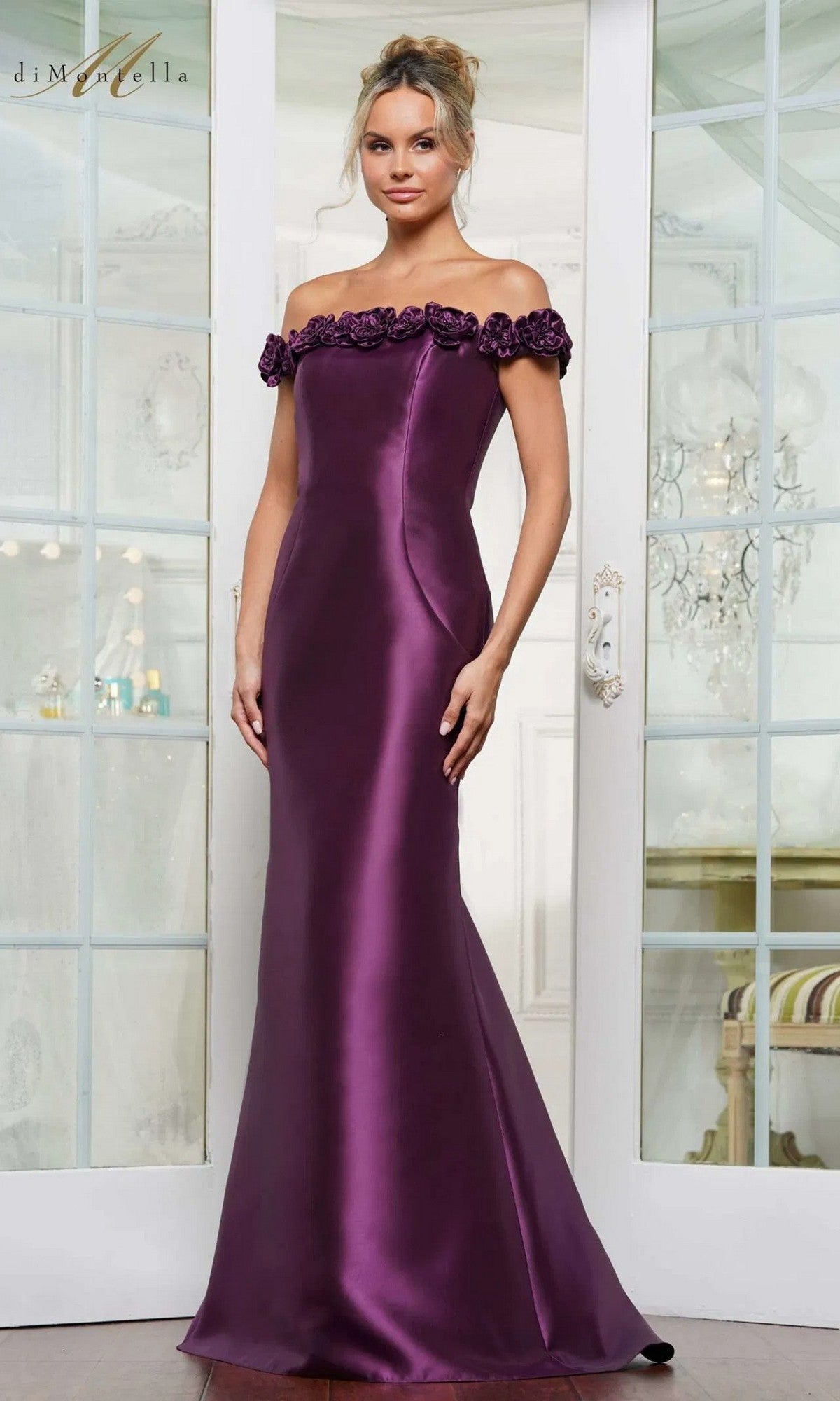 A woman stands indoors by glass-paneled doors, wearing the Rina Di Montella RD3205 long formal dress in grape—an off-the-shoulder, floor-length satin gown with floral neckline details and a graceful mermaid silhouette.
